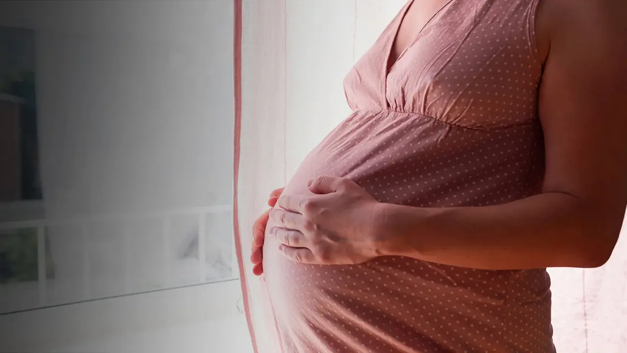 Pregnant woman in a pink dress cradling her belly by a window.
